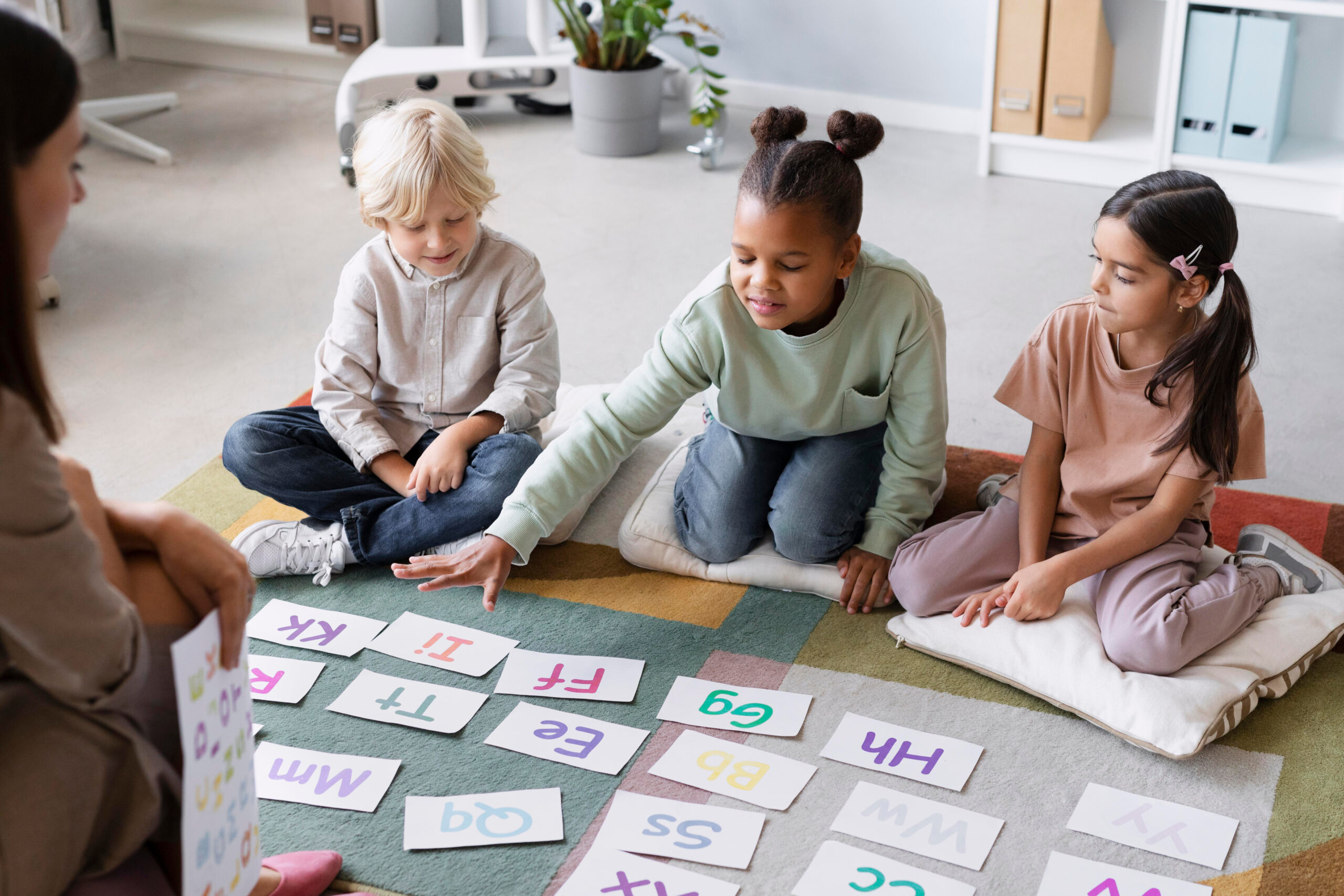 Three preschool-aged children sitting on a&nbsp;carpet in a&nbsp;classroom, with large letters in front of them as they learn.