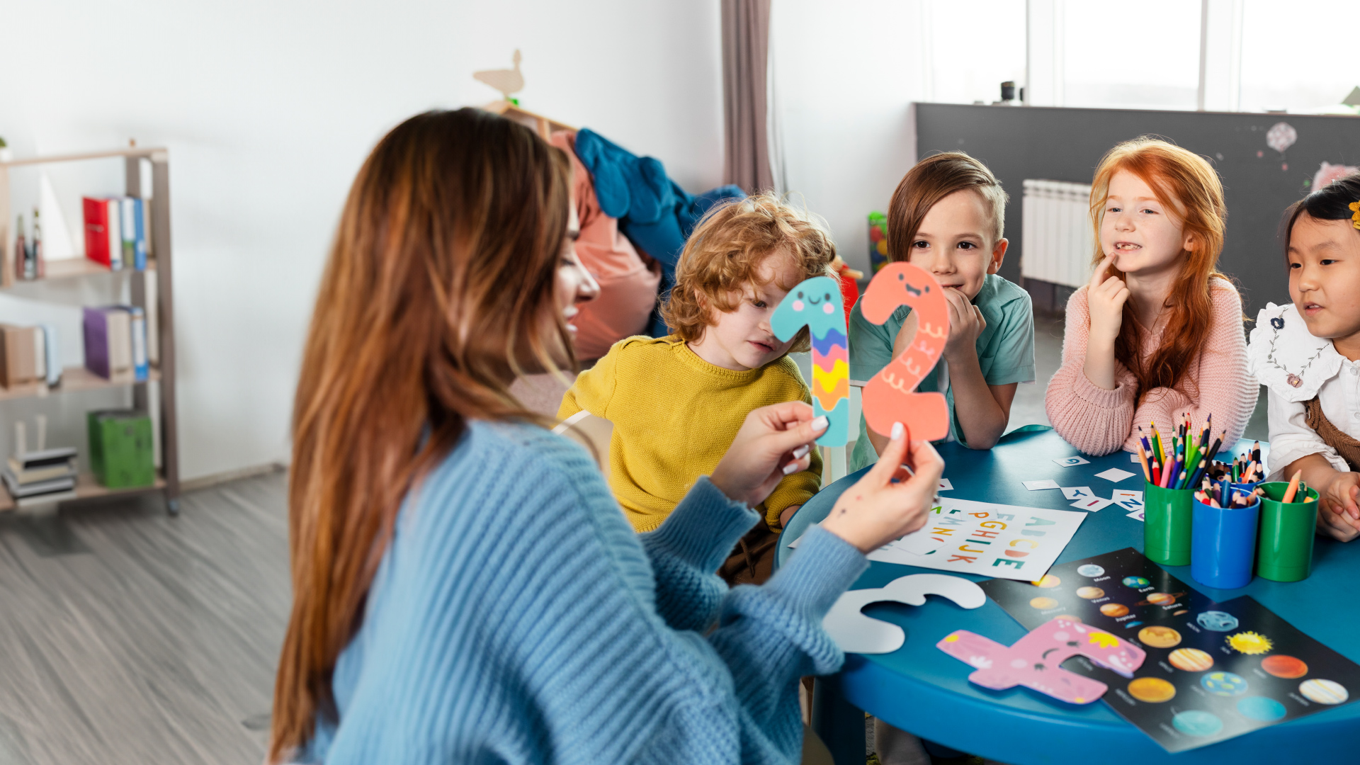 Children sitting at a table in kindergarten, looking at the numbers 1 and 2, held by the teacher leading the class