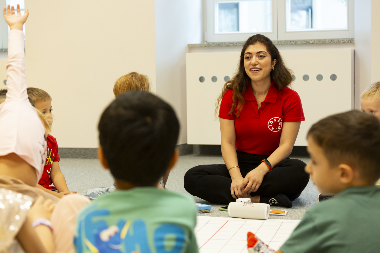 A preschool teacher sits cross-legged on a&nbsp;carpet in a&nbsp;red shirt, looking at a&nbsp;boy raising his hand while sitting in a&nbsp;circle with other children.