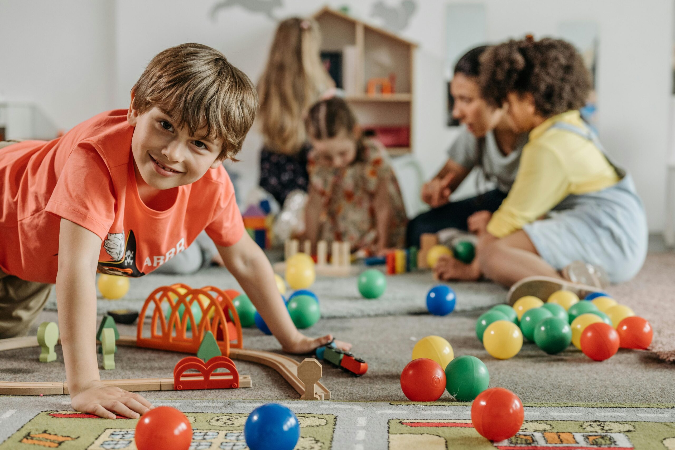A preschool-aged boy kneels on a carpet in preschool, looking directly at the camera, with other children playing in the background, toys, colorful balls, and a toy train track on the carpet.