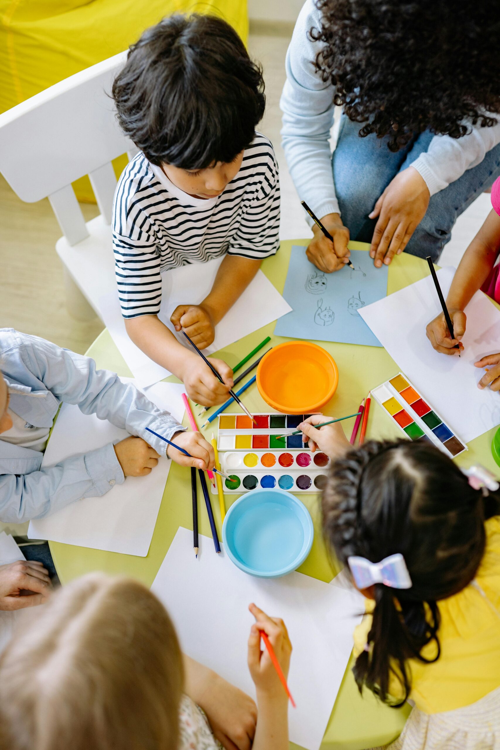Preschool children sitting at a table, painting, drawing, and writing with various tools, brushes, and crayons.