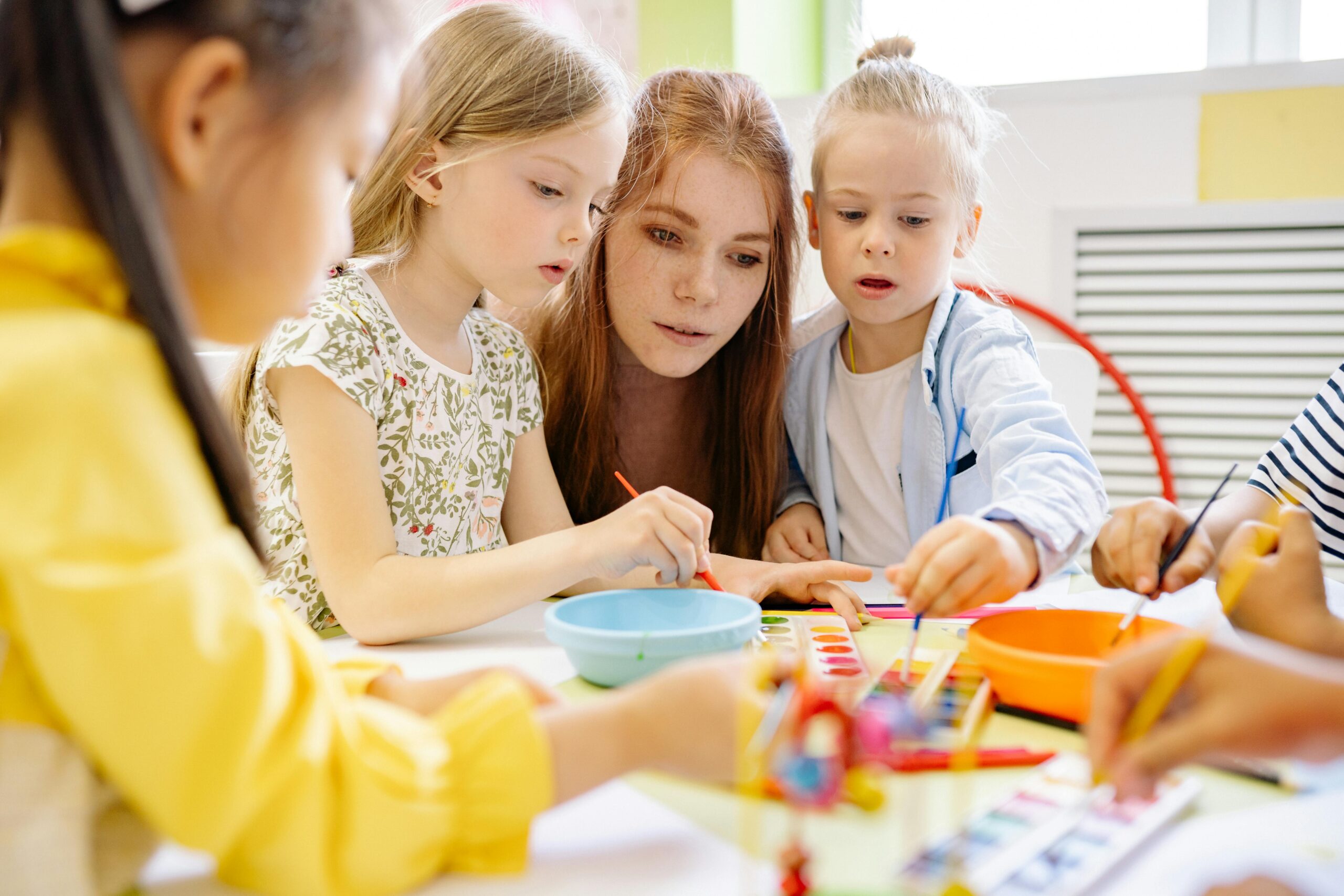 Two girls painting pictures with paints at a&nbsp;table in kindergarten, with a&nbsp;teacher sitting between them.