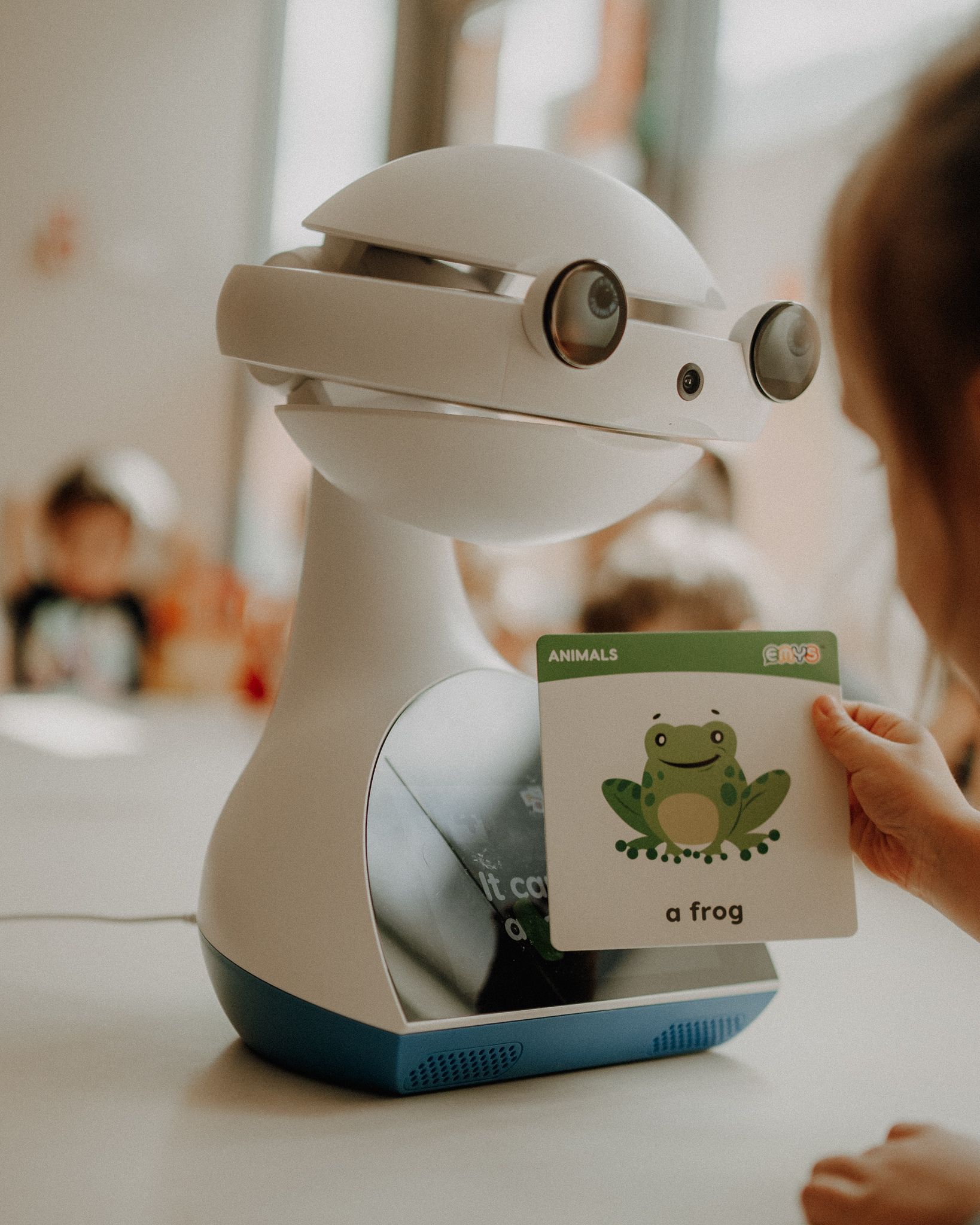 Robot edukacyjny Emys, który stoi na stoliku w przedszkolu, a dziecko przykłada do niego kartę z rysunkiem żaby i angielskim napisem frog.