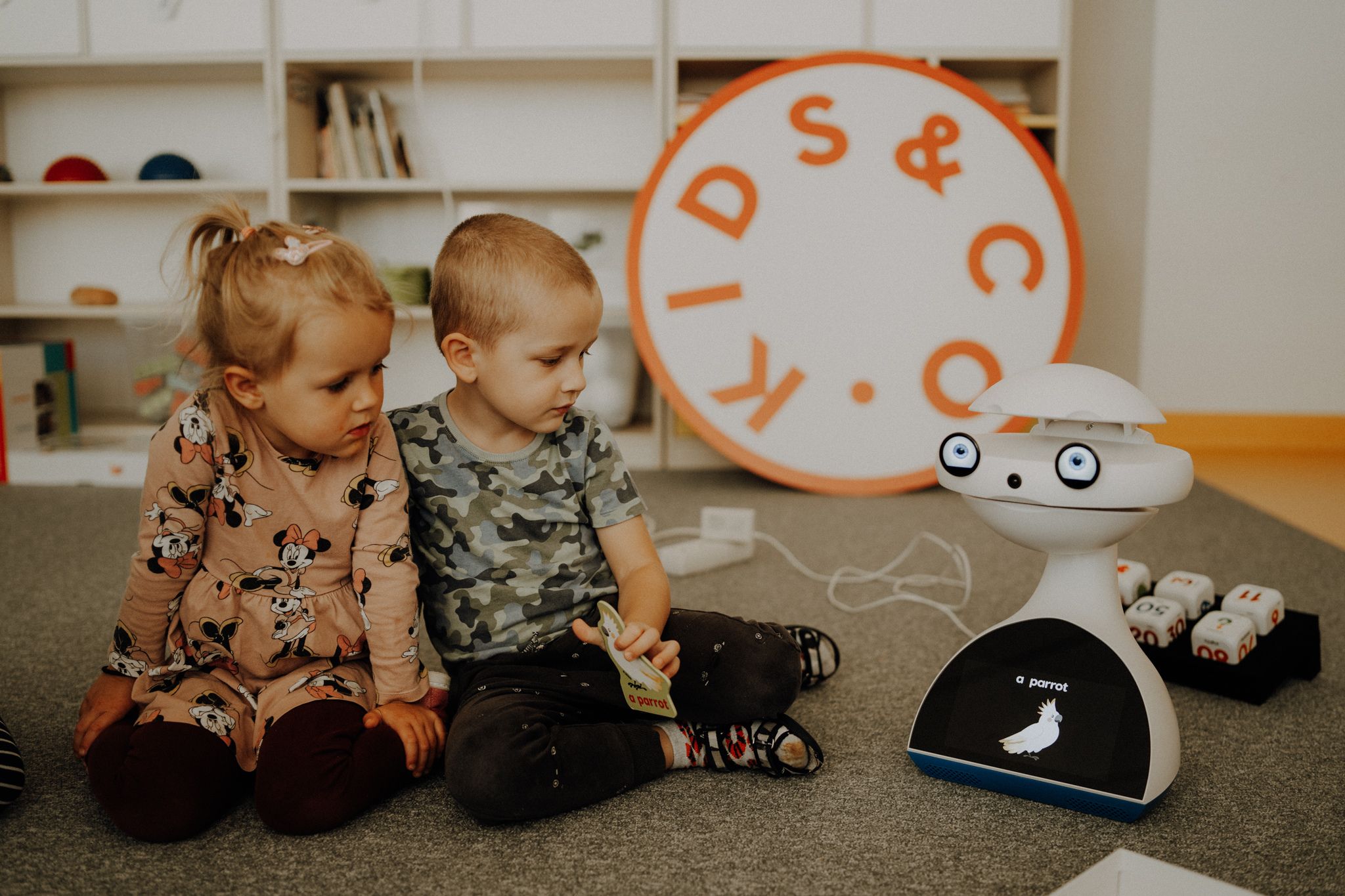 Dwójka dzieci w wieku przedszkolnym siedzi w sali przedszkolnej na dywanie i patrzy na robota do nauki Emys.