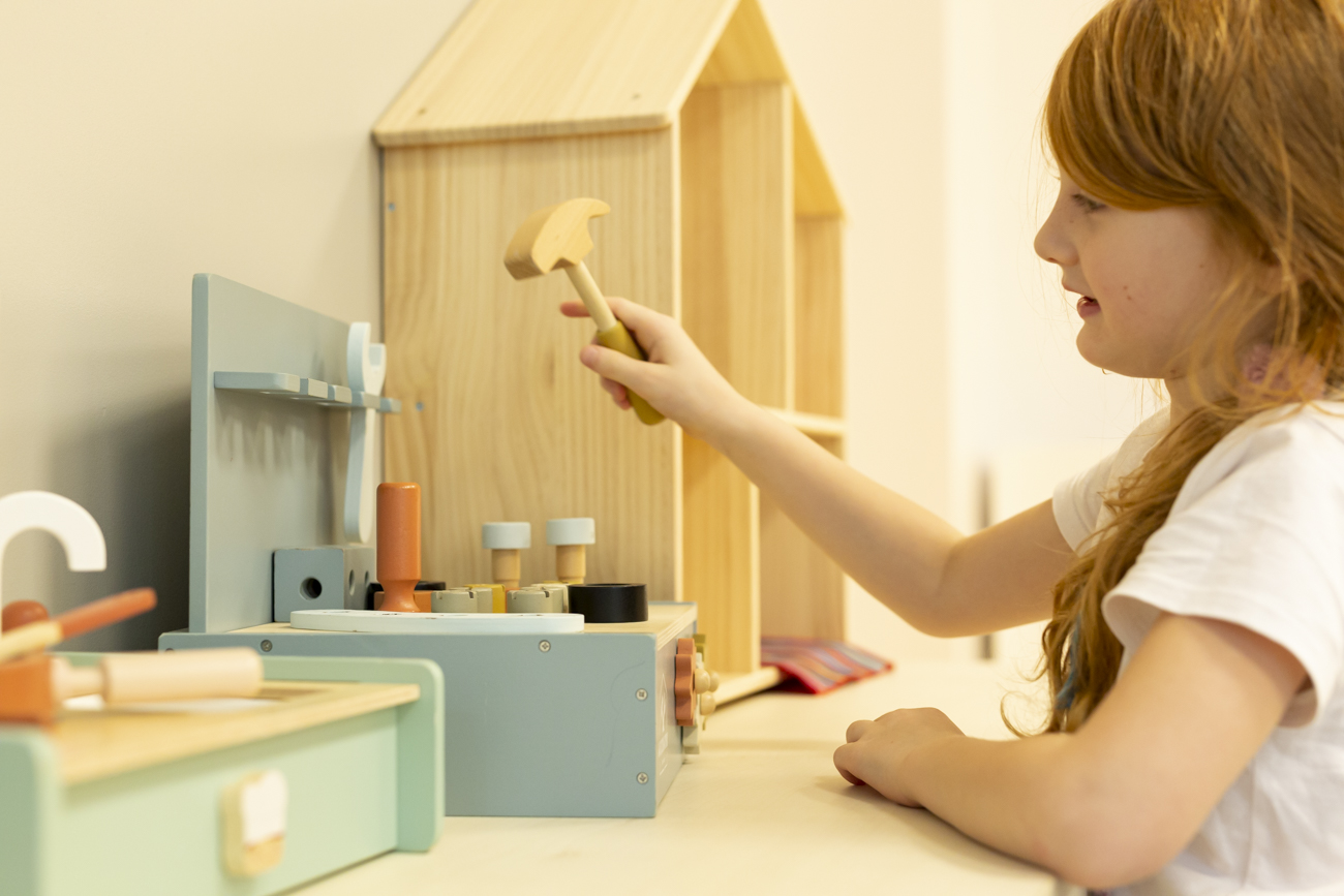 A preschool girl holds a toy hammer and is hammering toy nails into a base.