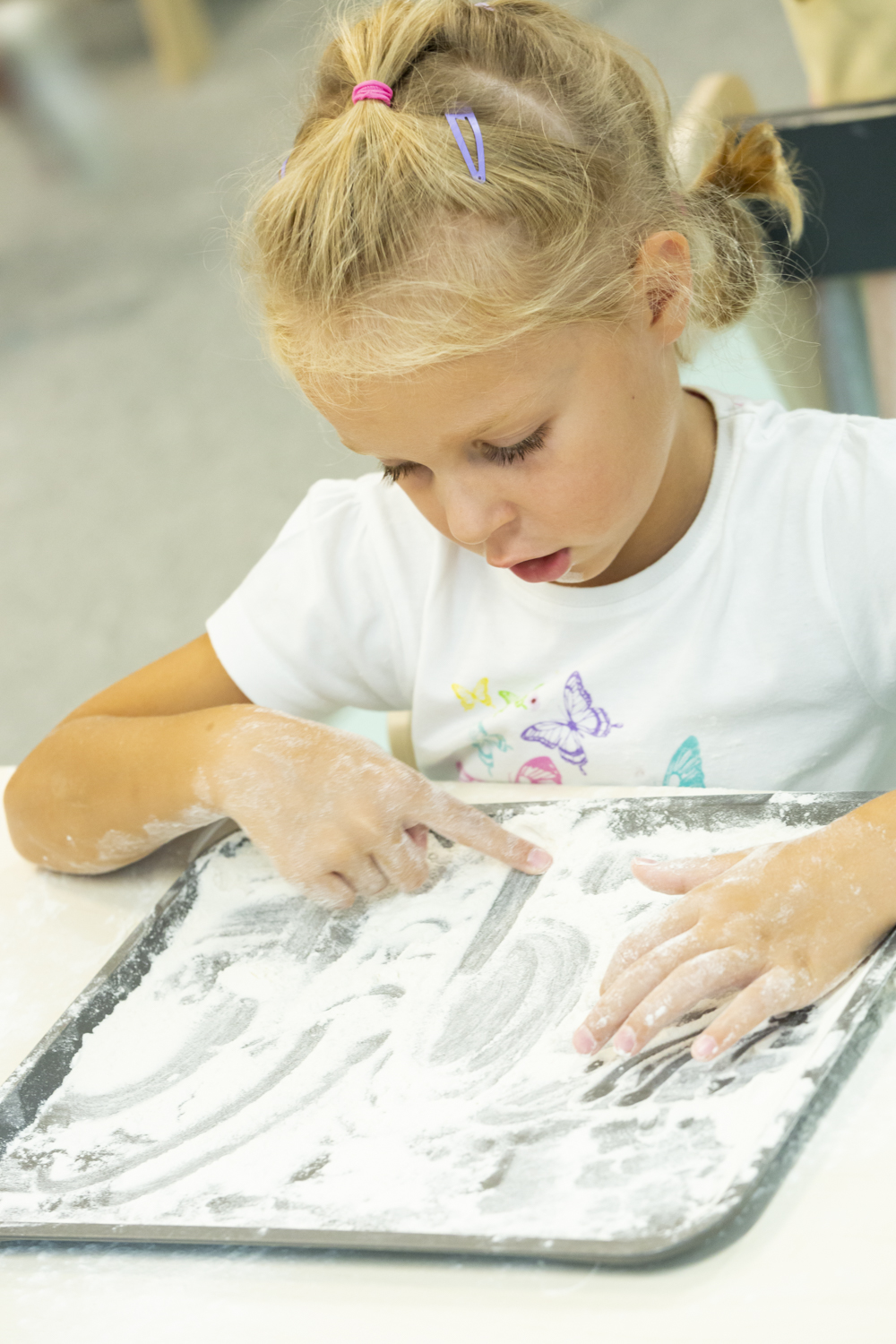 A preschool girl draws letters with her finger in flour.