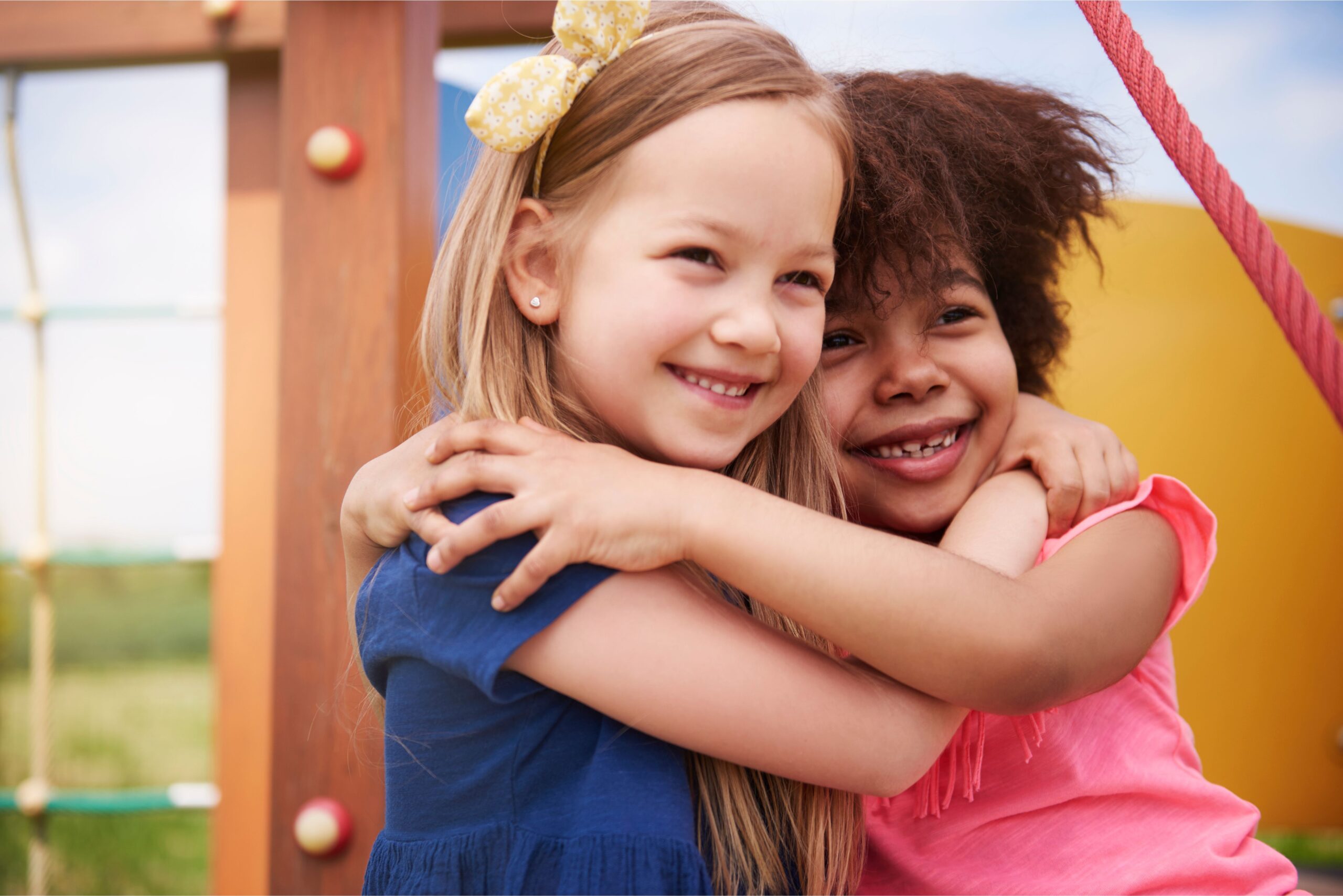 Two preschool-aged girls hugging and laughing – one blonde with light eyes, the other with dark skin and curly brown hair.