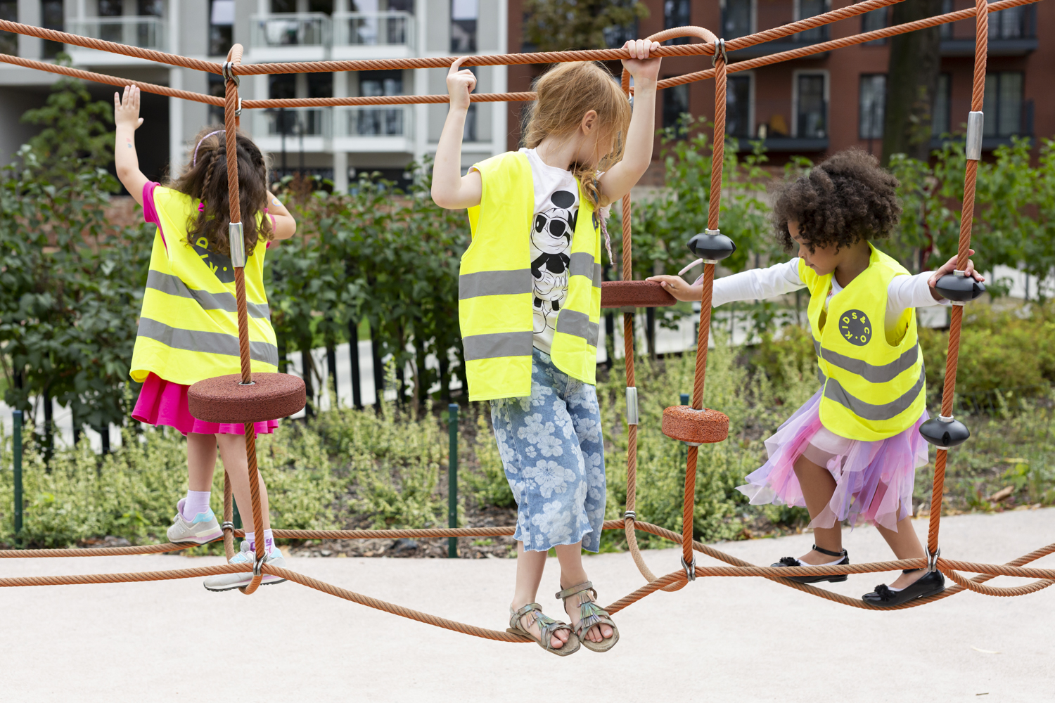 Three young girls of different skin colors, wearing reflective vests, playing on the playground, walking across a&nbsp;rope obstacle course.