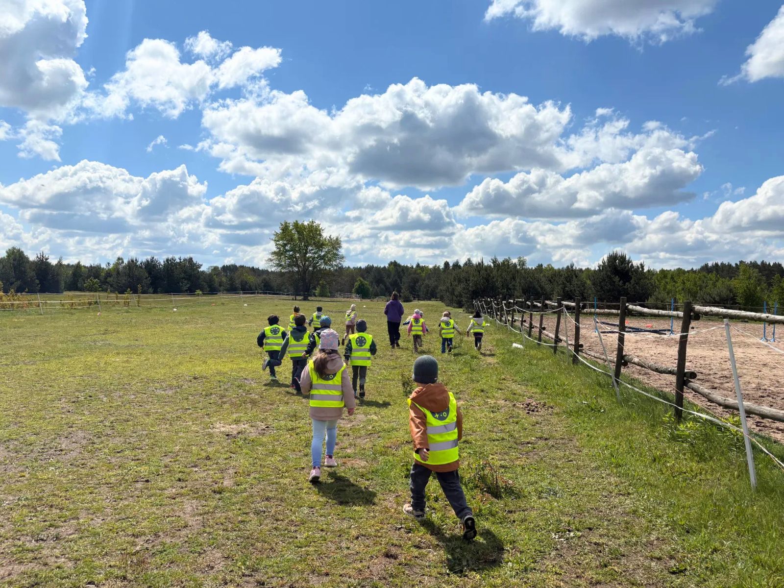 Preschool children wearing reflective vests running on the grass, their backs to the camera, with a&nbsp;forest and beautiful blue sky in the background.