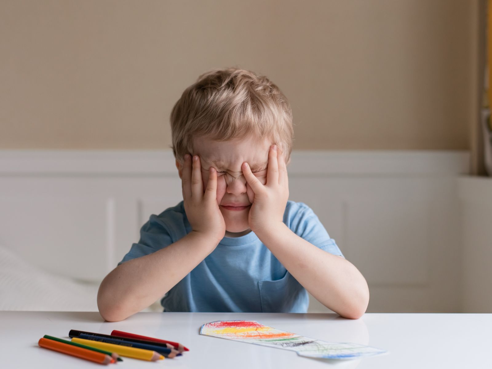 A young boy with blonde hair sitting at a&nbsp;table, covering his face with his hands, unable to concentrate, with colorful crayons laid out in front of him.