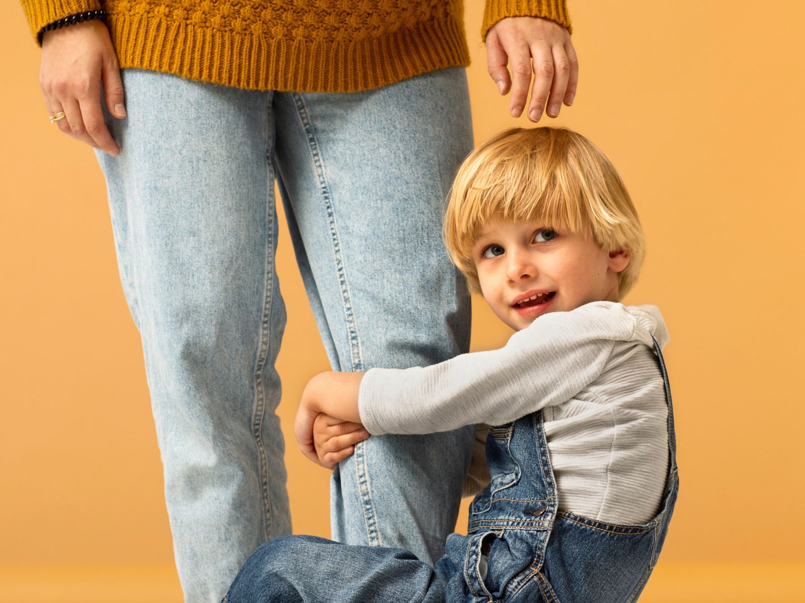 A preschool-aged boy with blonde hair clings tightly to his mother's leg, both standing against an orange background.
