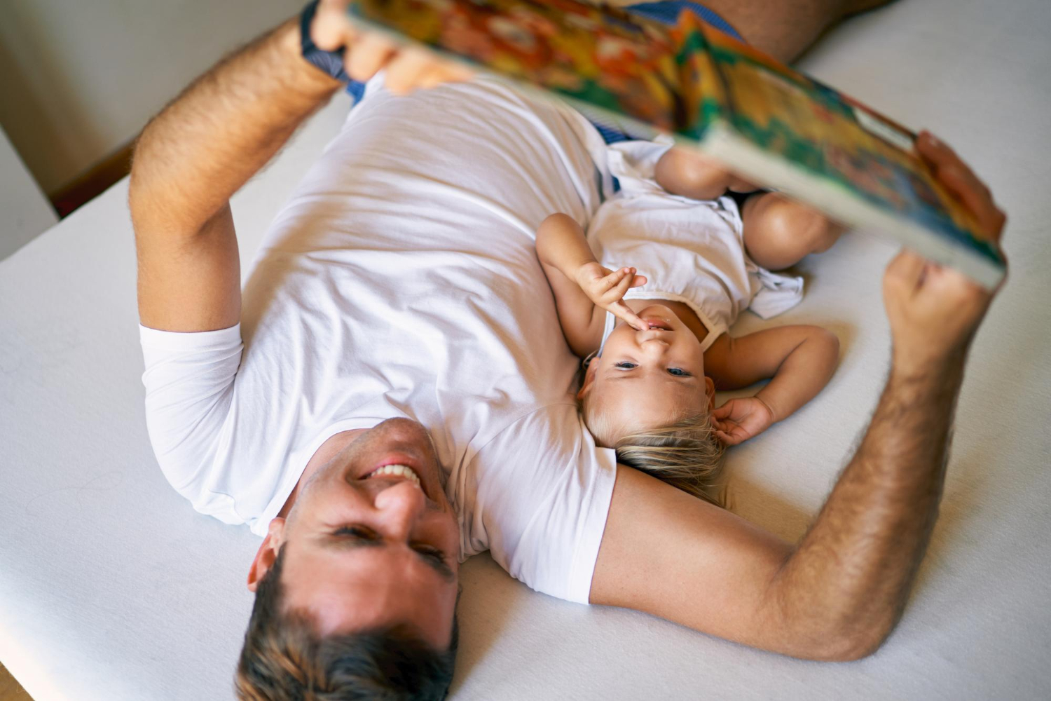 Dad and young child lying on a&nbsp;bed, dad holding a&nbsp;book above them as they read together.