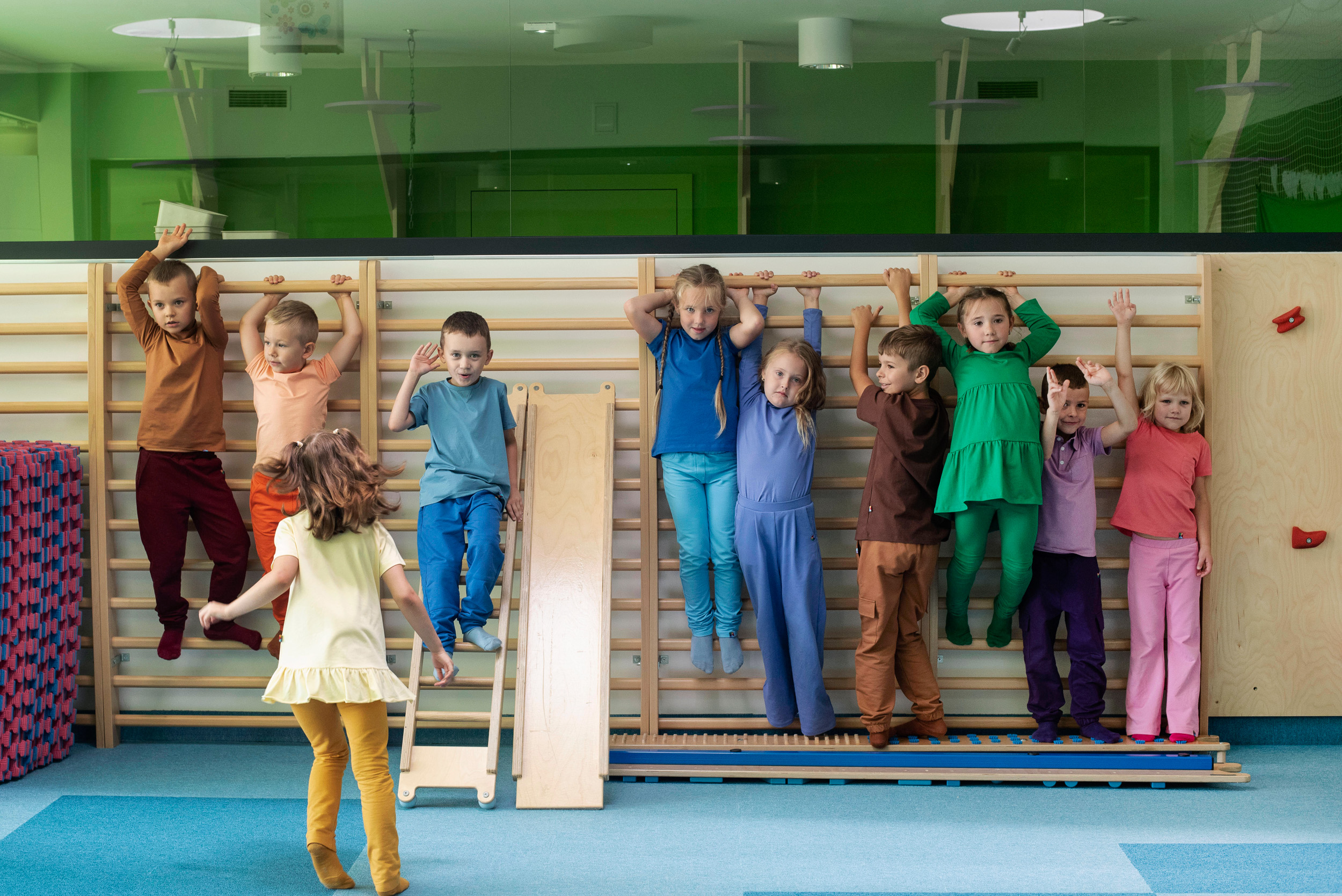 Children wearing colorful clothes exercise on wall bars in preschool.