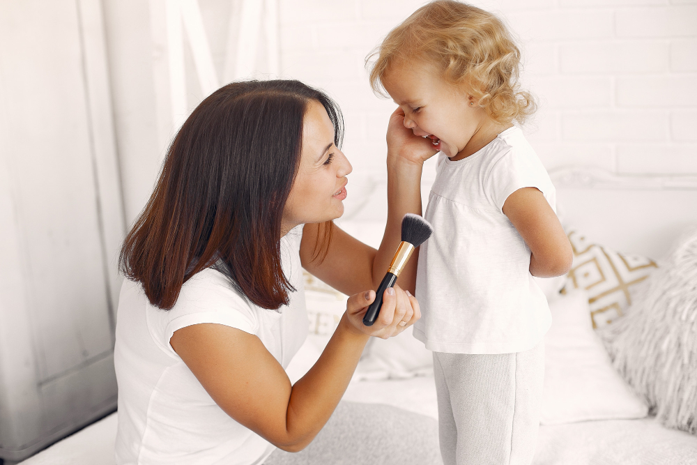Mama z&nbsp;małą córką uśmiechają się do siebie, mama trzyma w&nbsp;ręce pędzelek do malowania twarzy, obie są widoczne z&nbsp;profilu i&nbsp;ubrane w&nbsp;białe ubrania.